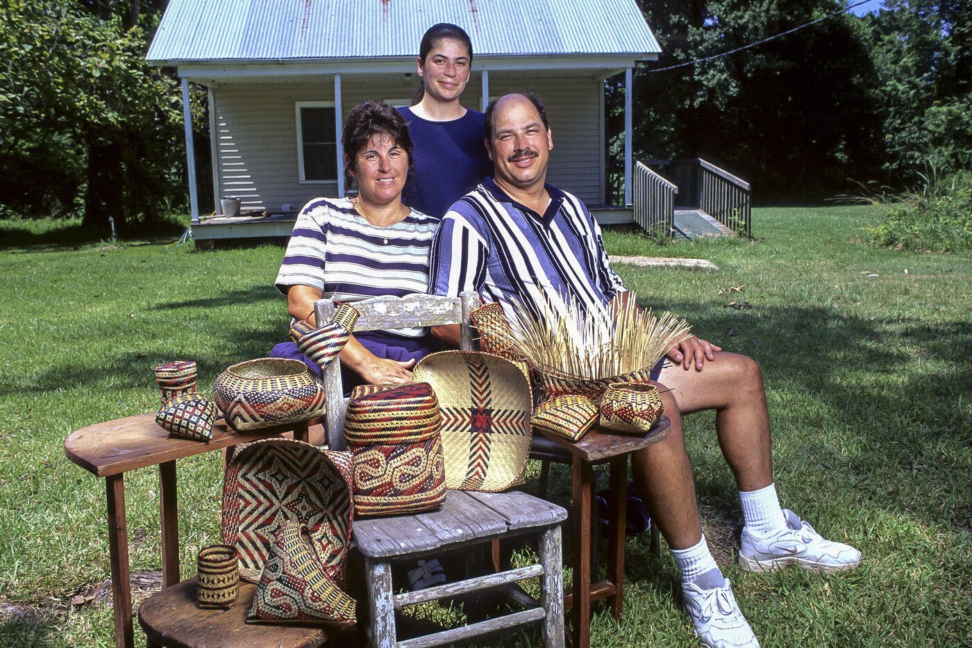 Three Chitimacha Triberibal members, one standing and two seated with some of their baskets on wooden furniture in front of a small house.