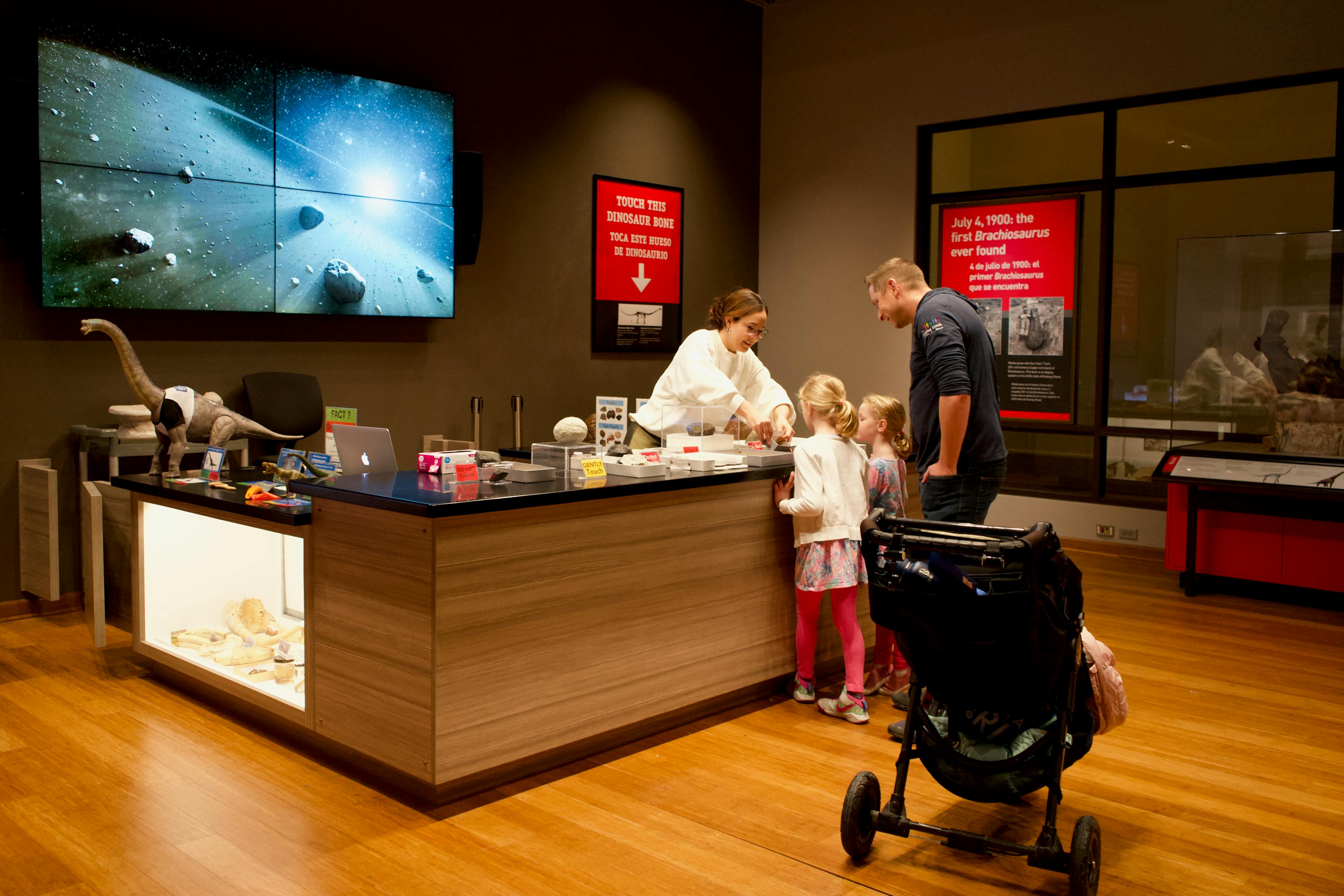 An adult and two children interact with a museum staff member who stands behind a counter in the Science Hub. On the counter are objects and specimens related to the content in the exhibit. A large video screen hangs on the wall behind the counter.