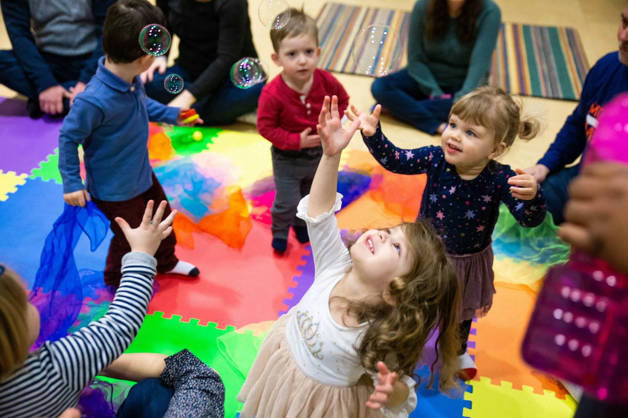 Five young children reach for bubbles above them. The floor is covered in soft, colorful tiles.