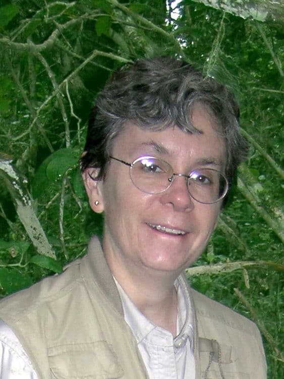 A women with short gray hair and glasses smiles in front of a leafy forest backdrop