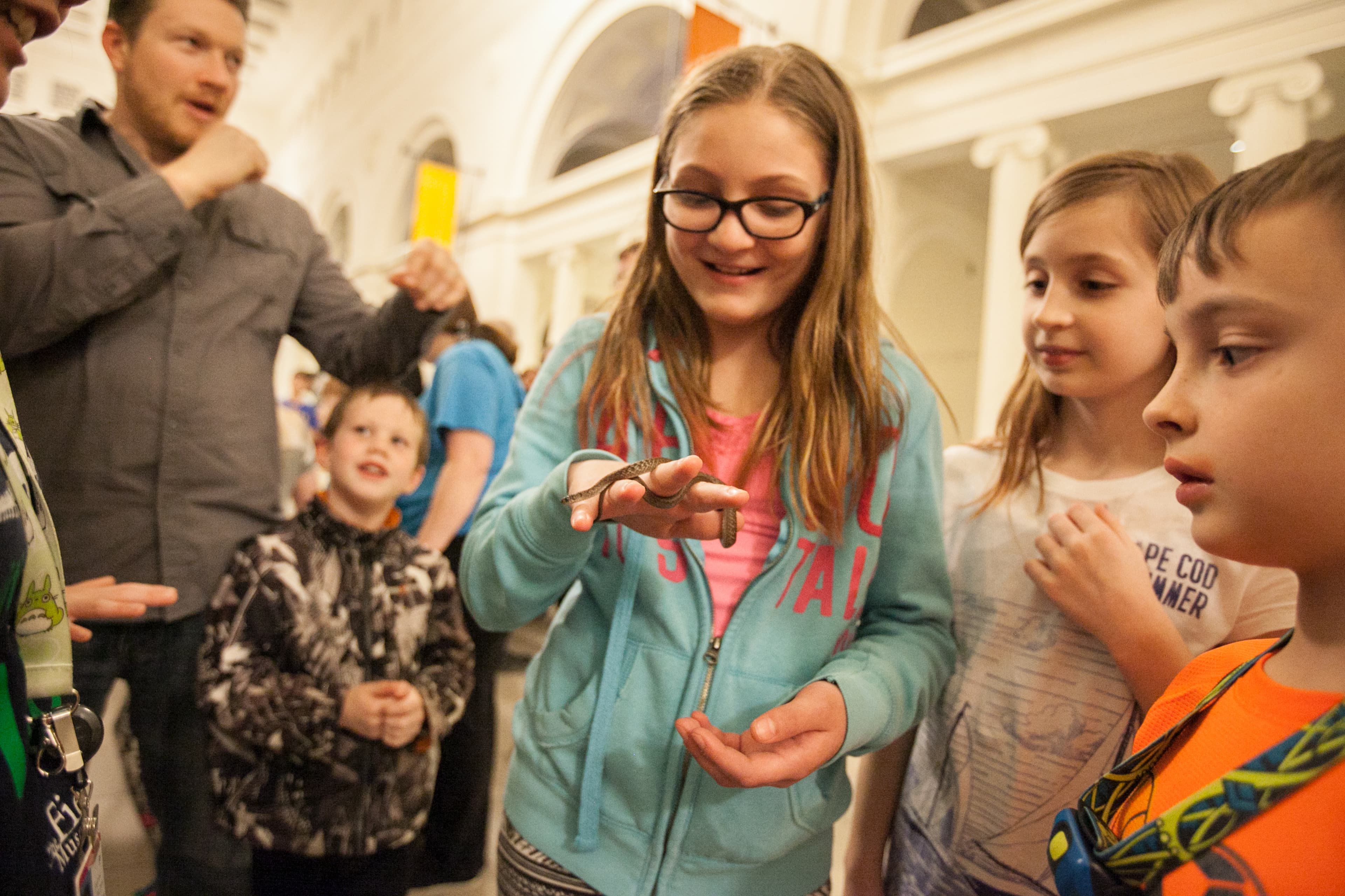 A young visitor holds a baby snake in one hand while other visitors look on.