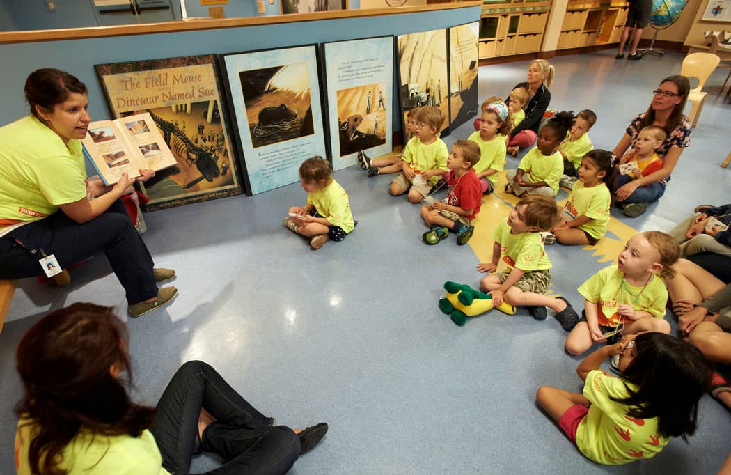 A group of young children sit on the floor listening to a woman read to them. Most everyone in the photo are wearing matching bright yellow t-shirts.