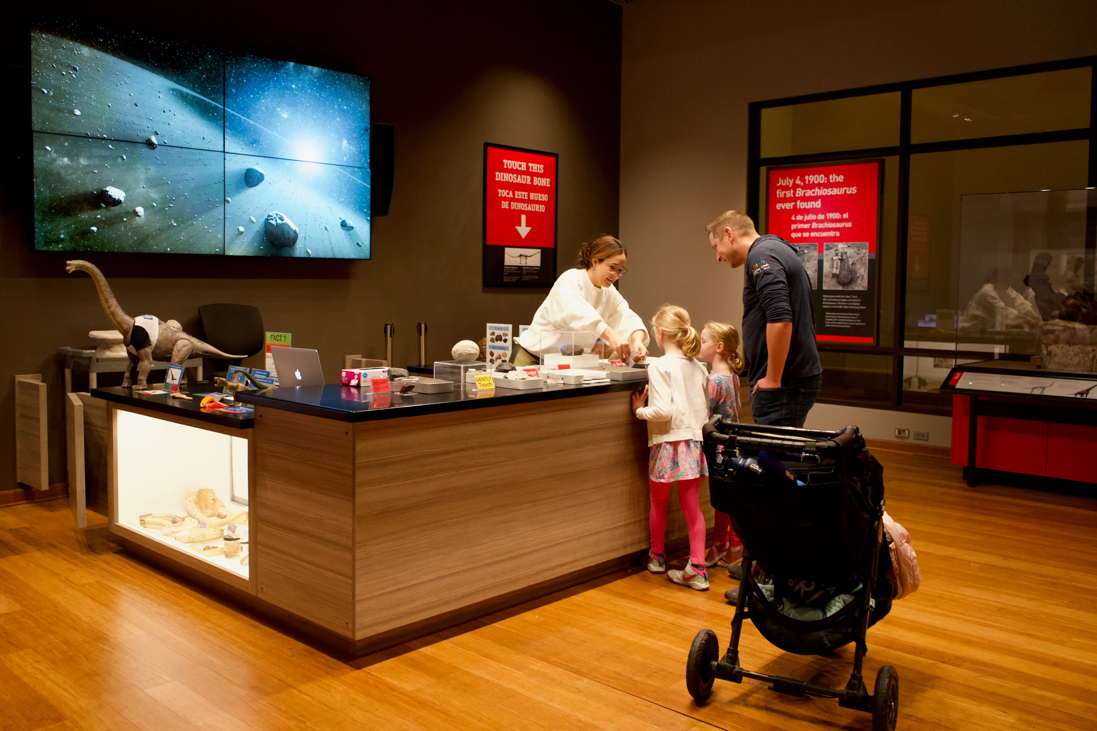 An adult and two children interact with a museum staff member who stands behind a counter in the Science Hub. On the counter are objects and specimens related to the content in the exhibit. A large video screen hangs on the wall behind the counter.