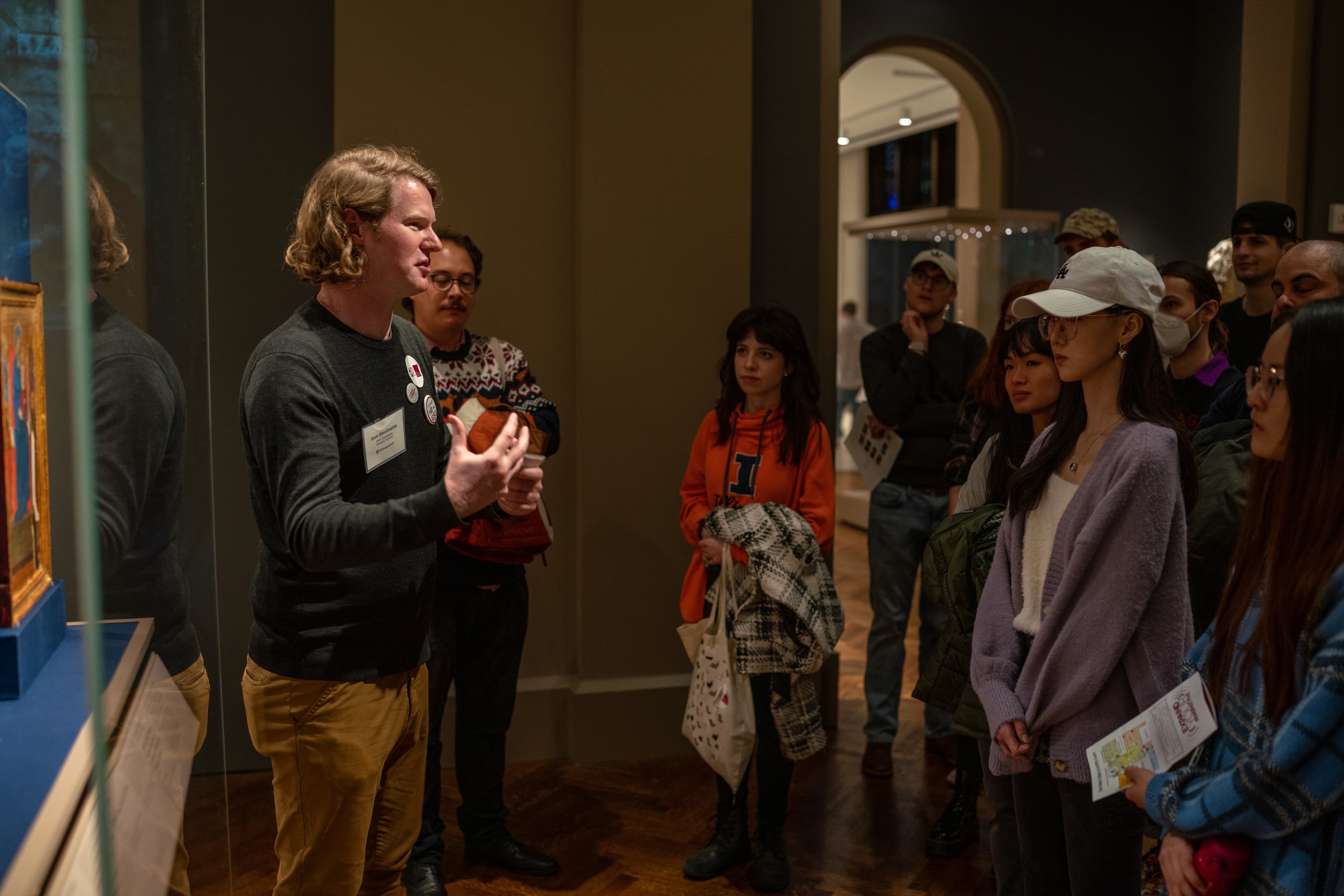 A group of students stands facing a person speaking in a exhibition