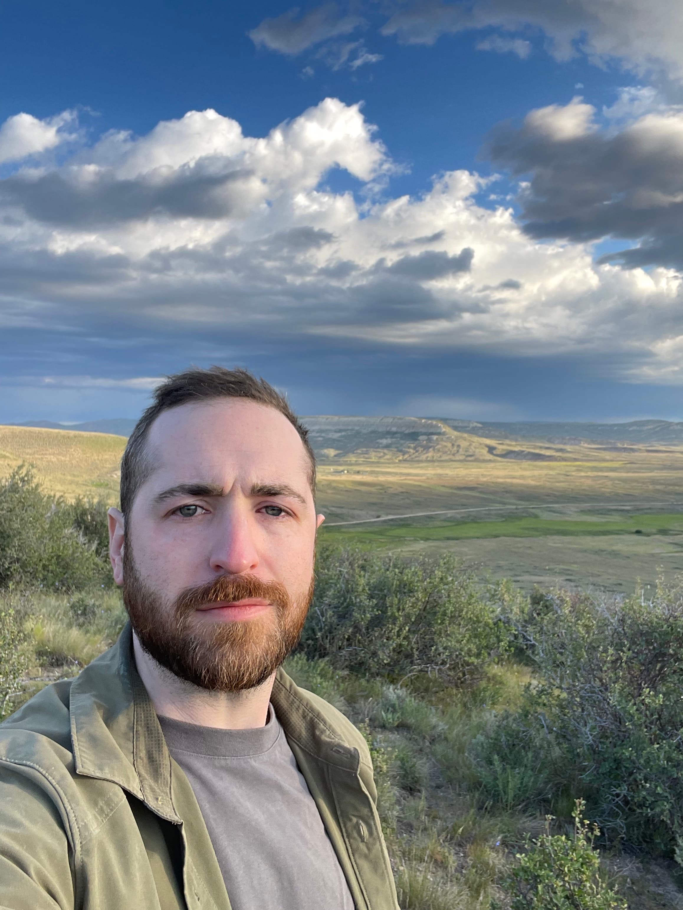 A man stands in front of an open landscape with rolling grassy hills. White and grey clouds fill the blue sky.
