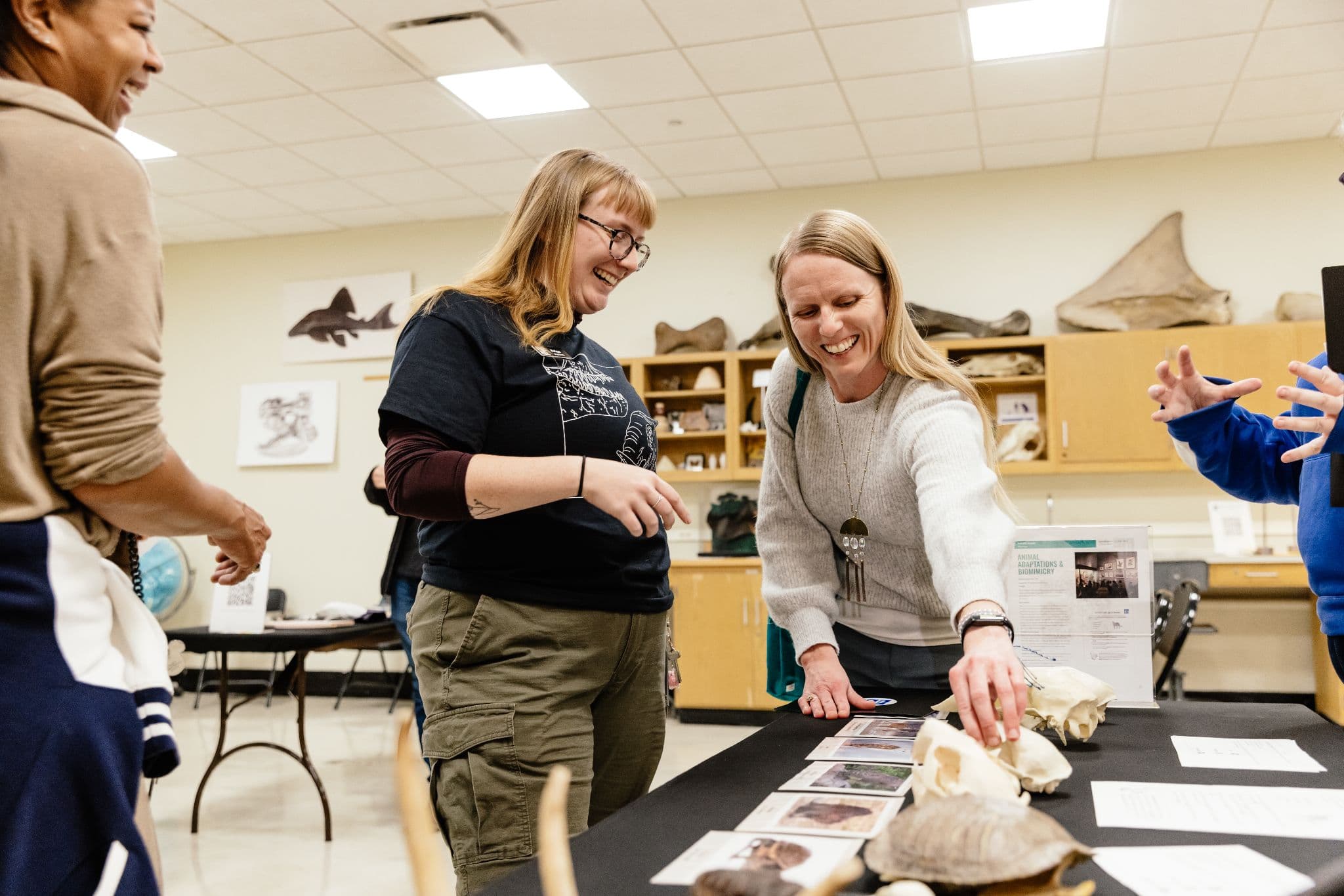 Two woman smile as they look down at objects on a table. Two other people can be partially seen to each side.