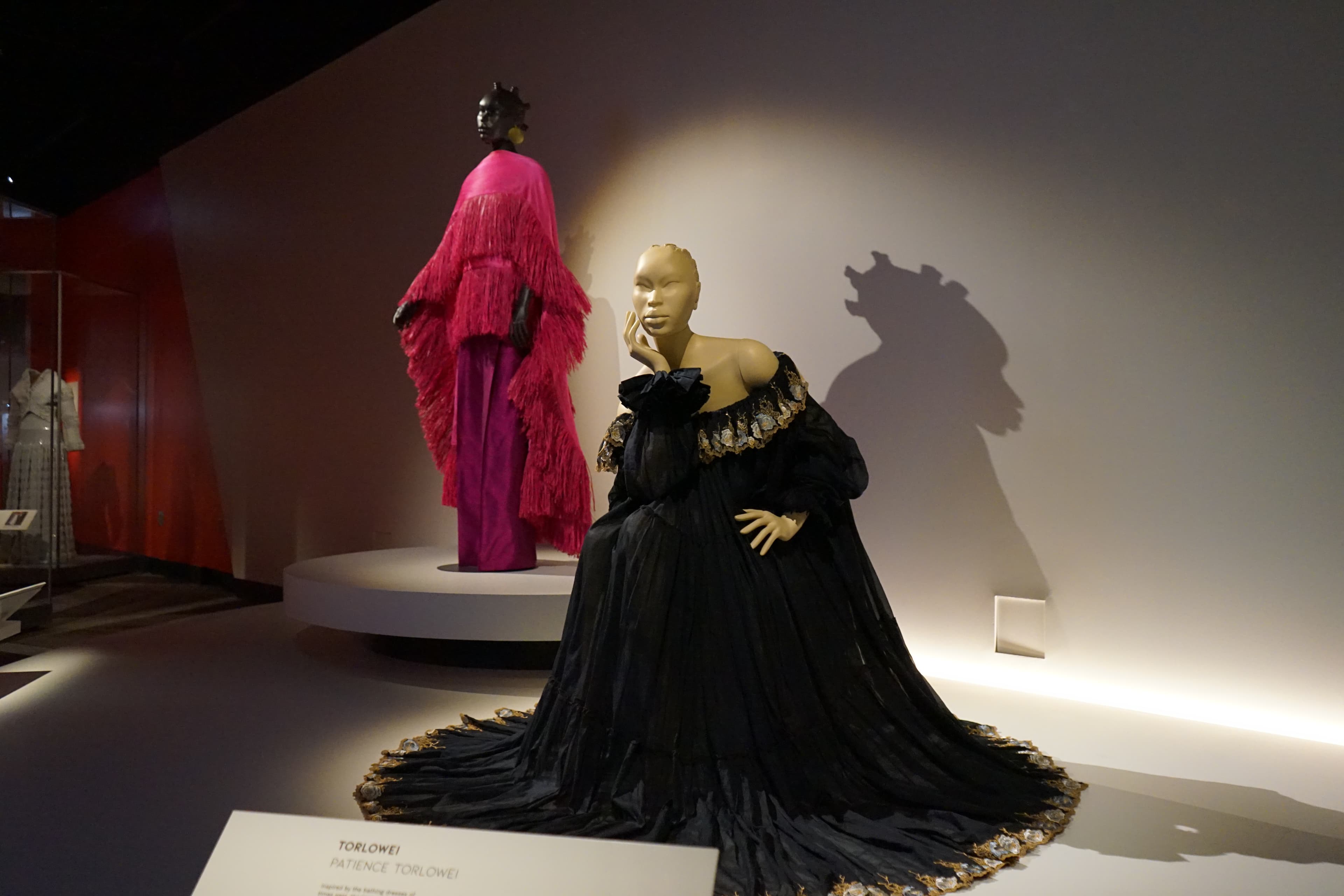 A black dress on a seated mannequin, with another mannequin standing in the background wearing a bright pink dress.