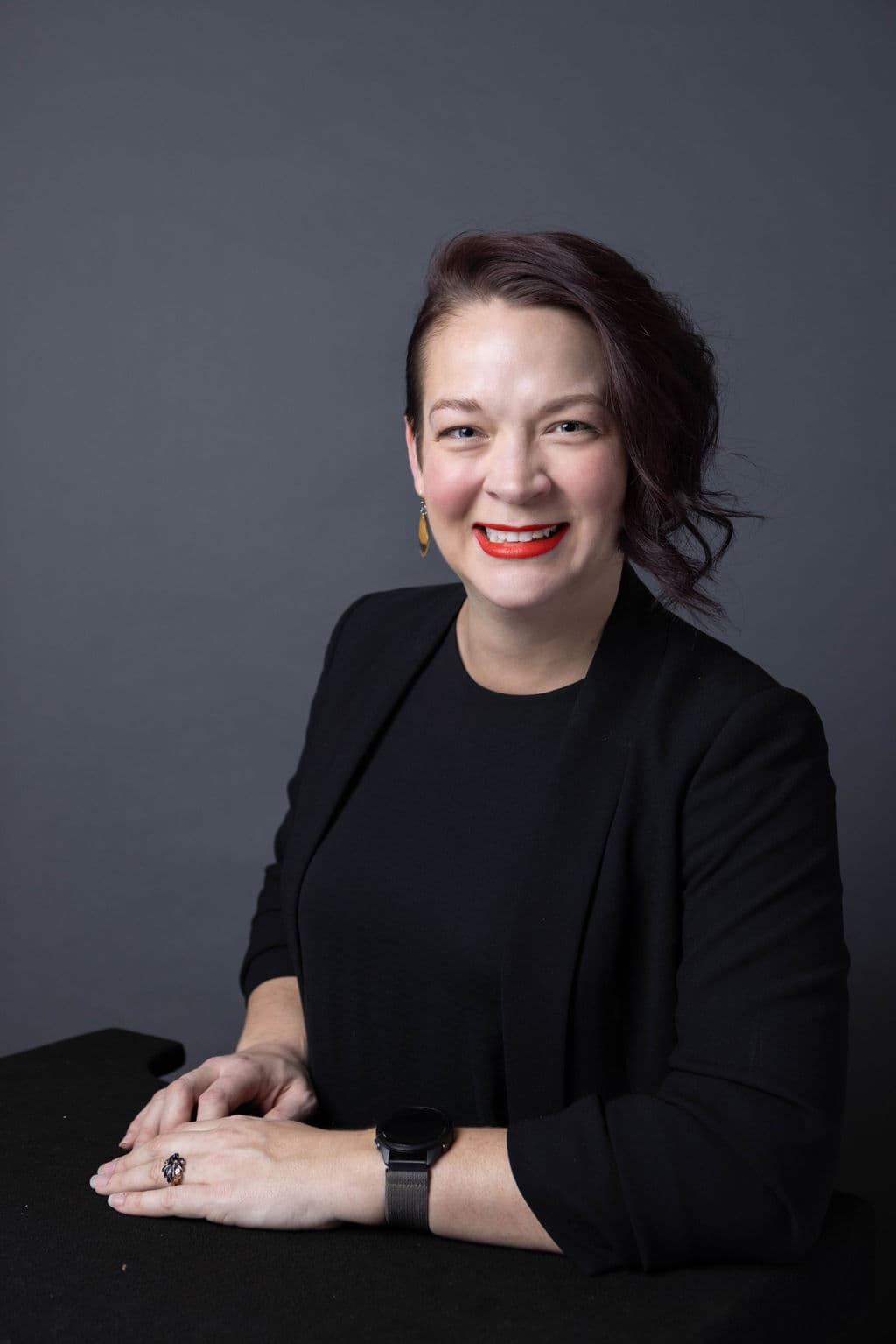 A woman wearing a black shirt and jacket, with her arms resting on a table in front of her.