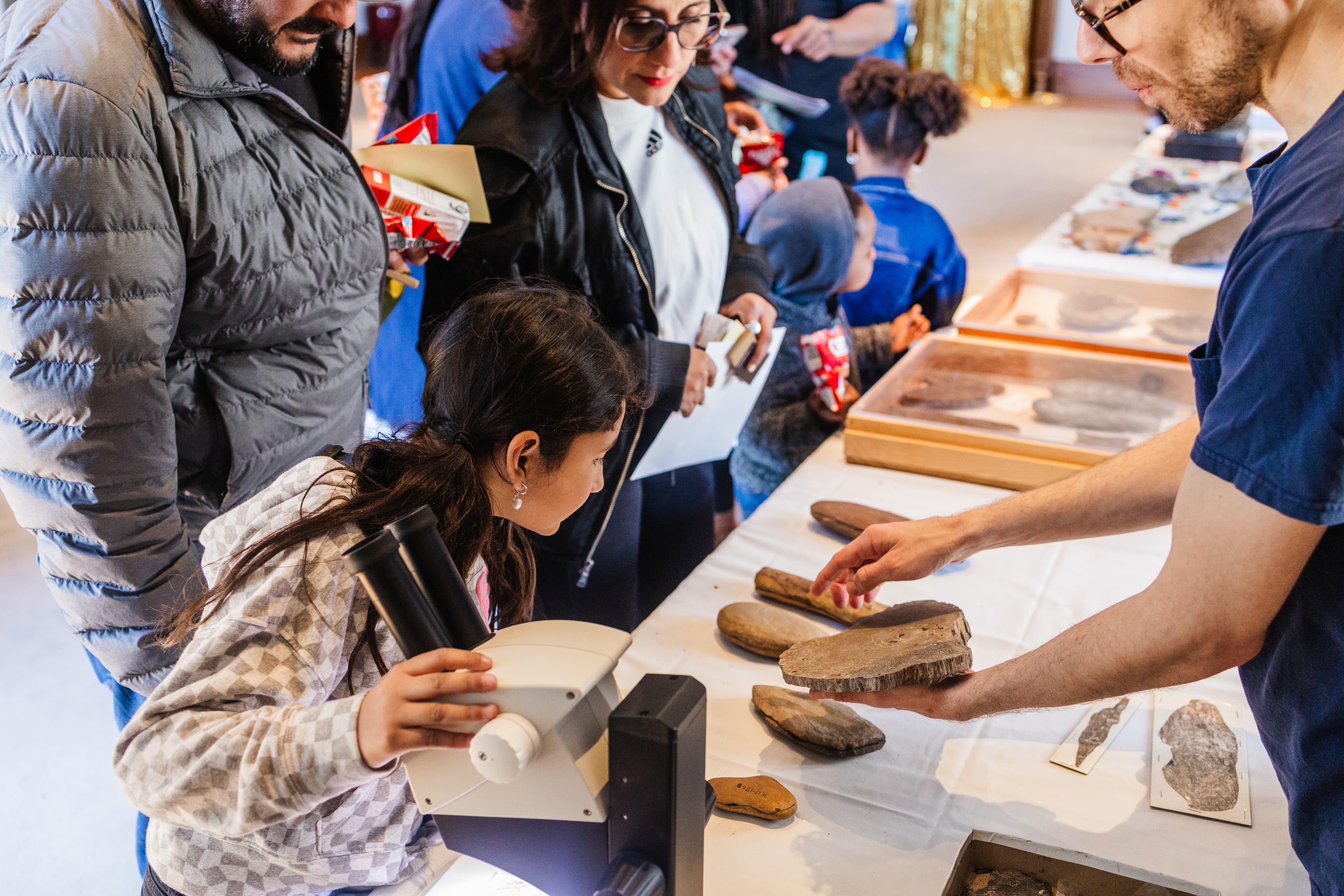 Adults and children at a table filled with museum specimens, some in glass cases. A staff member is holding one object out for the group to see more closely