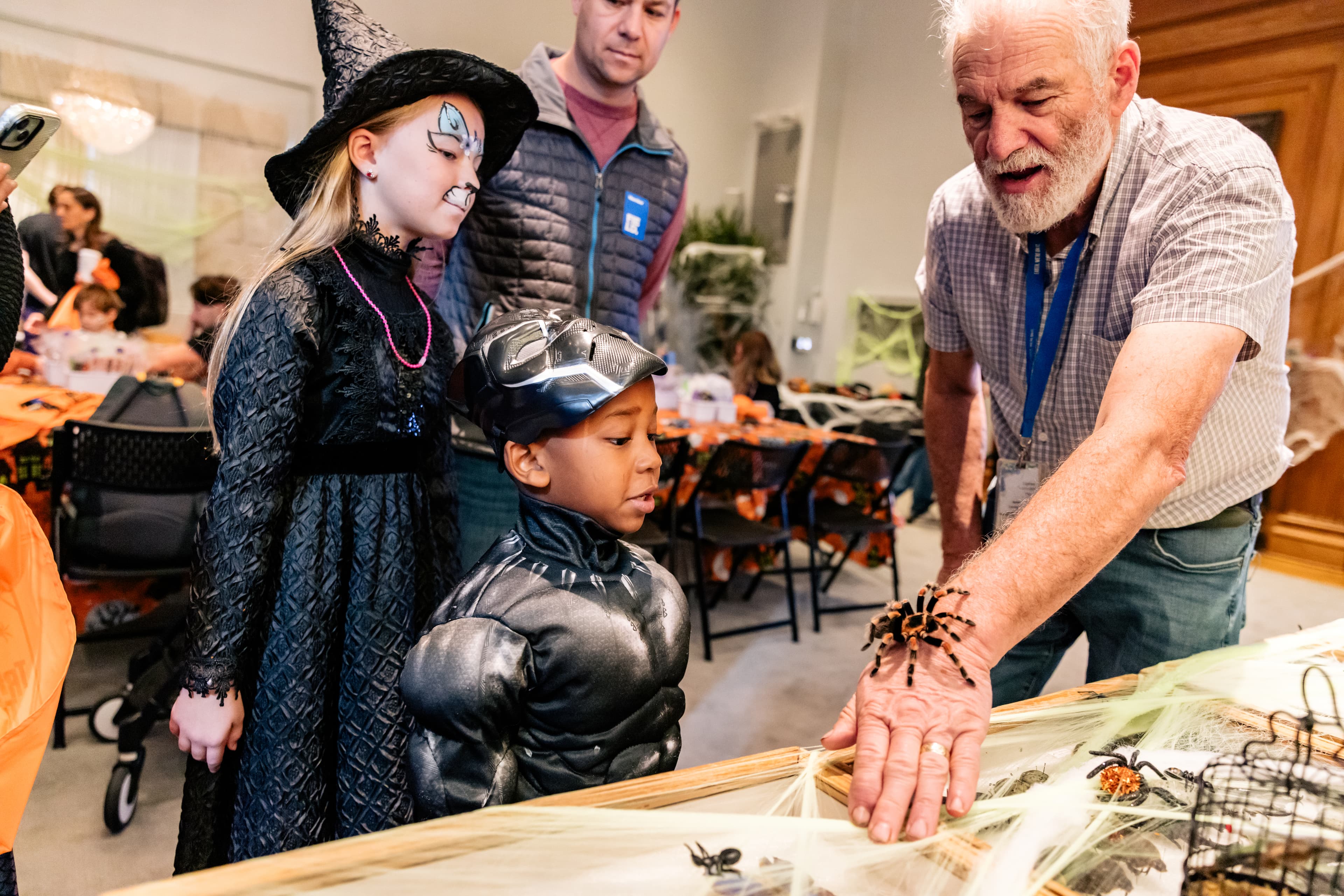 Museum staff member extends his arm as a tarantula crawls on his wrist. Museum visitors in halloween costumes look on.