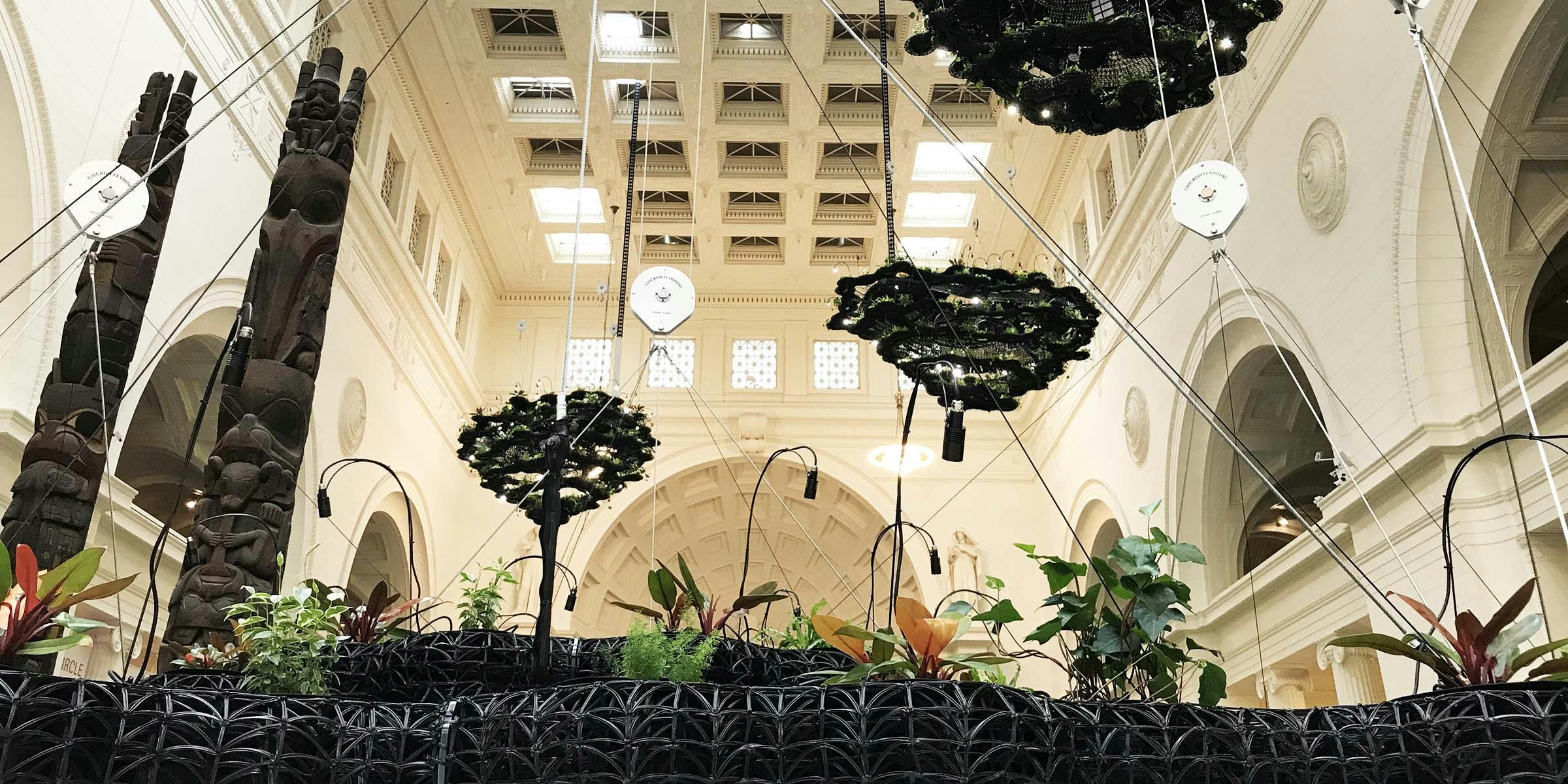 In the foreground, an eye-level view of a black plastic structure holding plants. The background includes the museum's Stanley Field Hall, with two totem poles to the left and three cloud-like black plastic structures suspended from the ceiling.