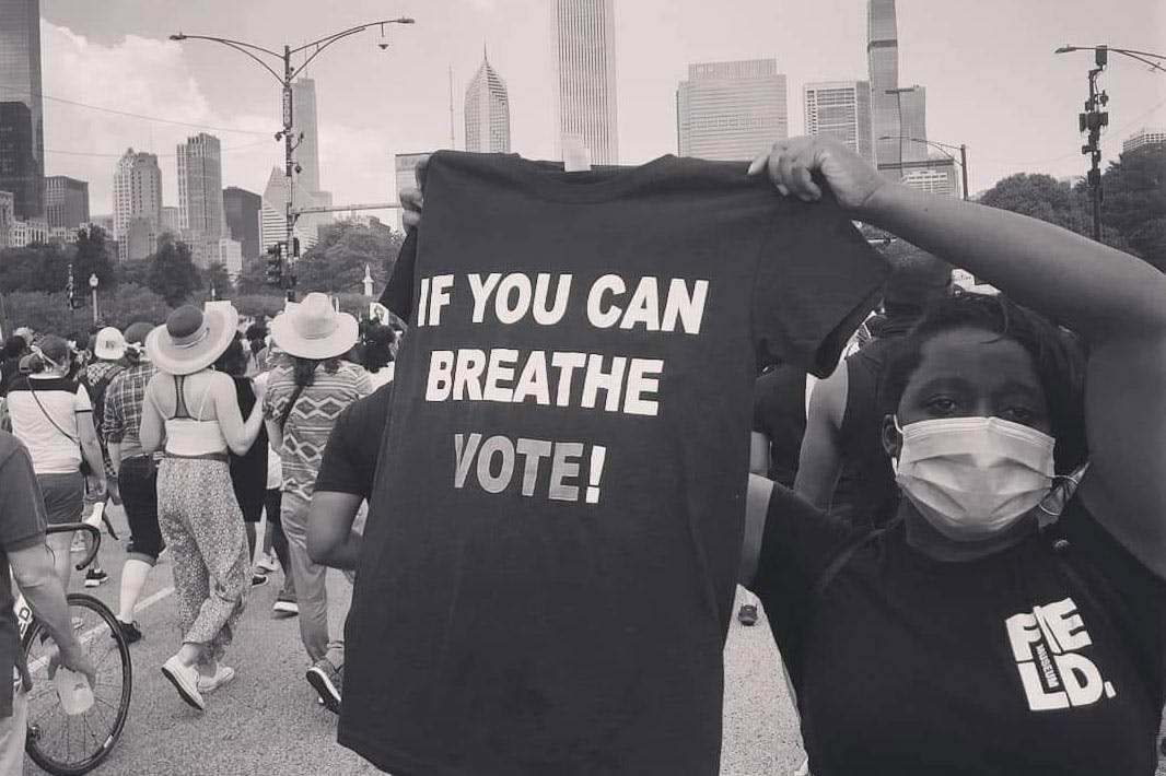 Ylanda Wilhite at a protest in downtown Chicago. She wears a facemask and a Field Museum shirt while holding up a t-shirt that reads, “If you can breathe, vote!”