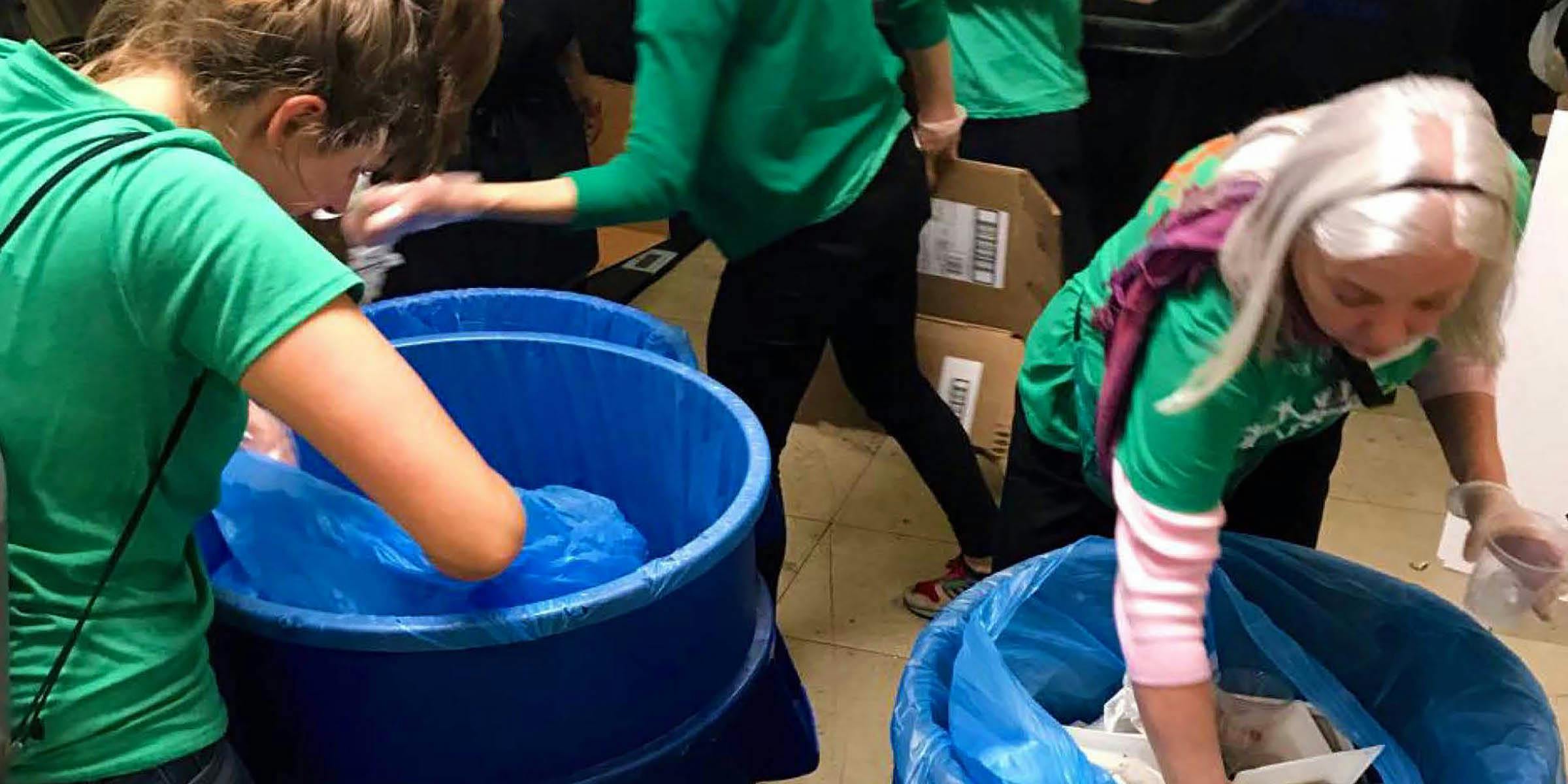 Two women wearing green shirts reaching into large blue recycling bins