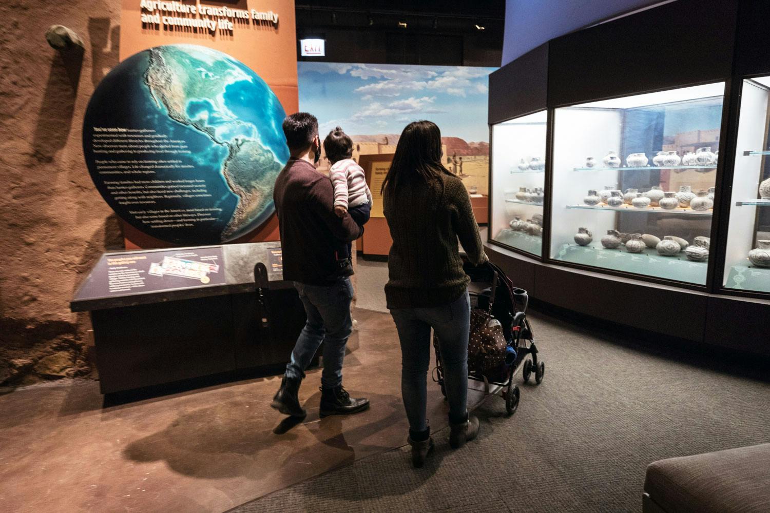 Two adults and a small child walk through the Ancient Americas exhibition. A map of the globe is on the wall to the left, and many pots are in a display case to the right.