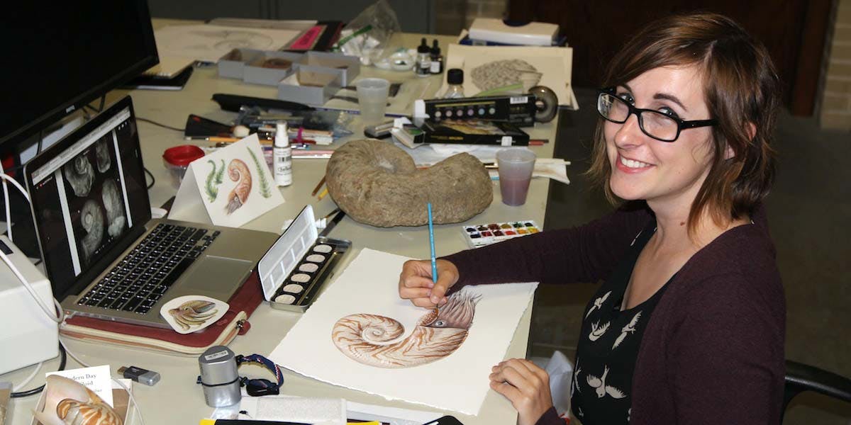 A woman sitting at a desk with art supplies, holding a pencil in hand and drawing a nautilus
