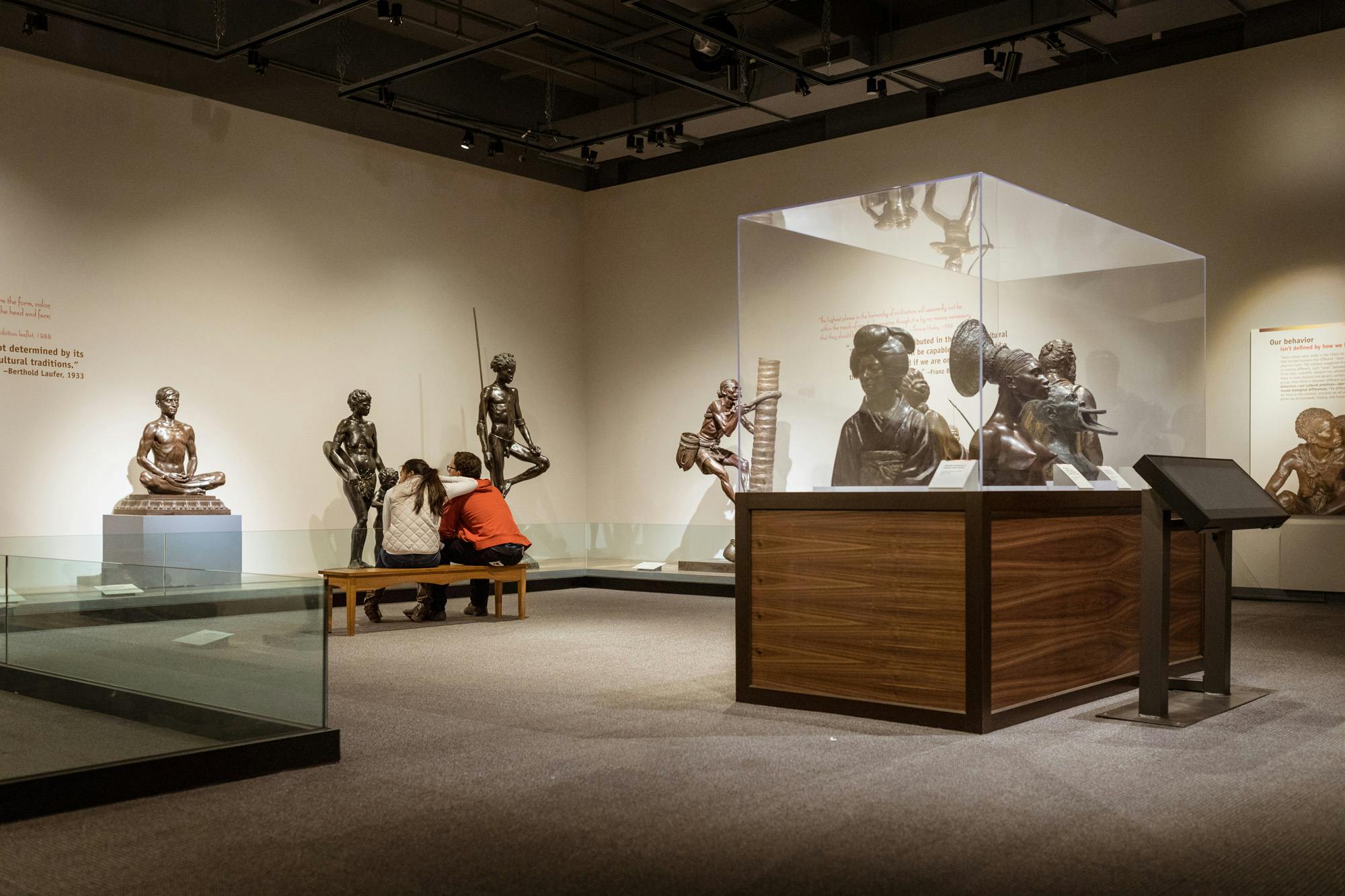 Two visitors sit together on a bench, looking at life-size sculptures of people by Malvina Hoffman. In the foreground several bronze busts are displayed in a glass case.