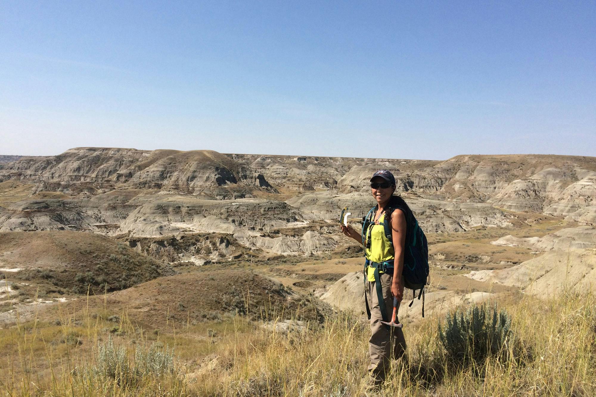 Akiko Shinya holds a pick hammer and stands in a grassy area with badlands in the background.