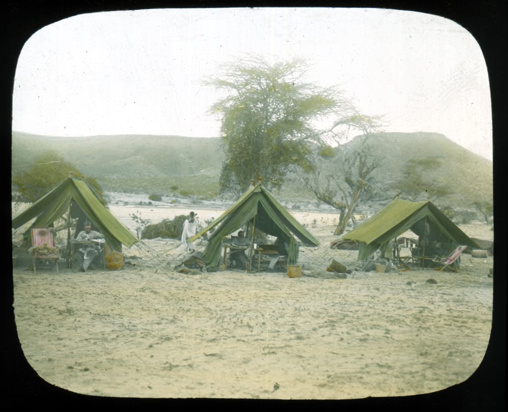 A colored photograph with black border, showing three tents in a row. The foreground is sandy, and the background has a few sparse trees and mountains.