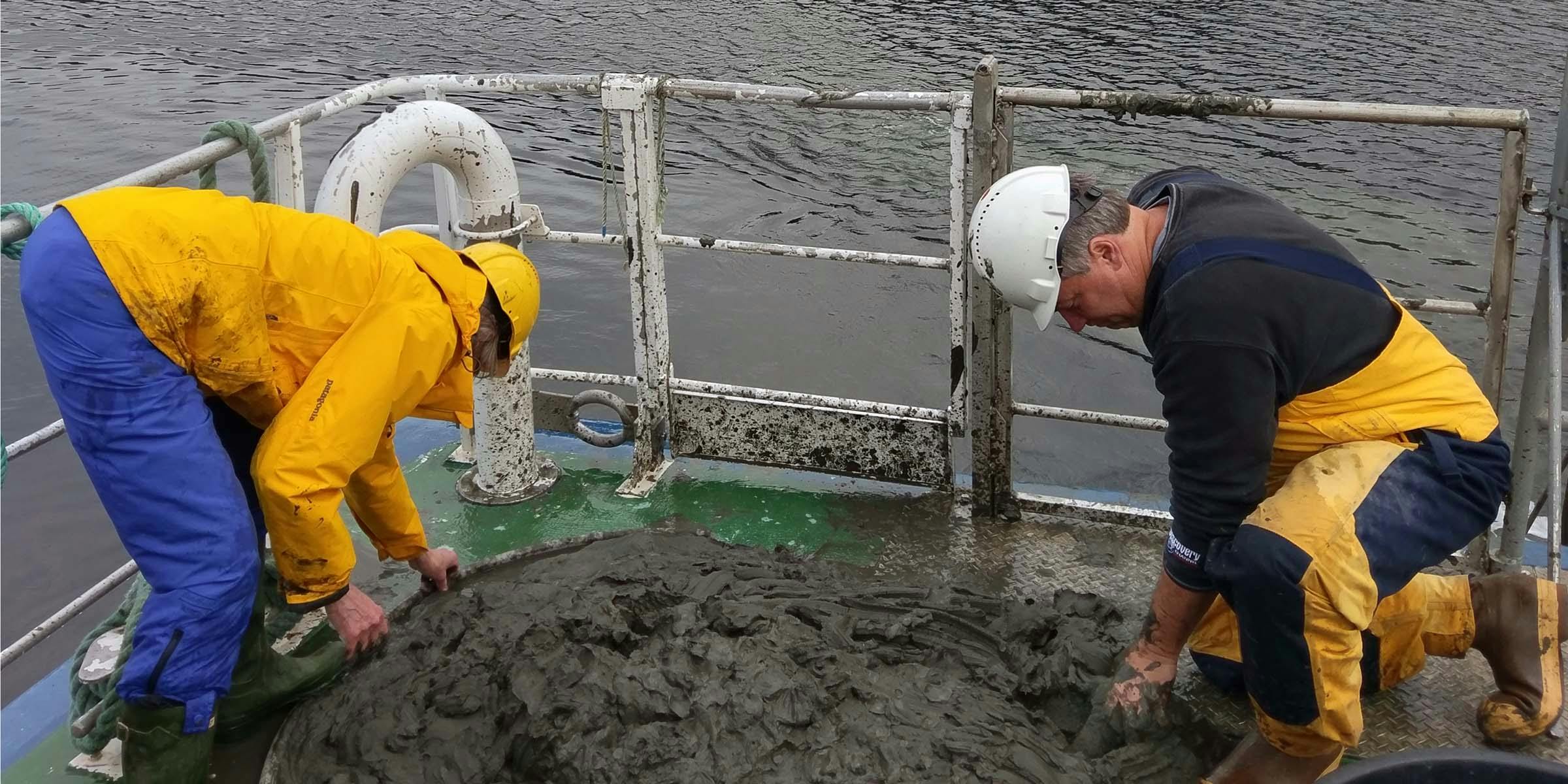 Two people wearing hard hats on a boat, leaning over and examining mud on the deck