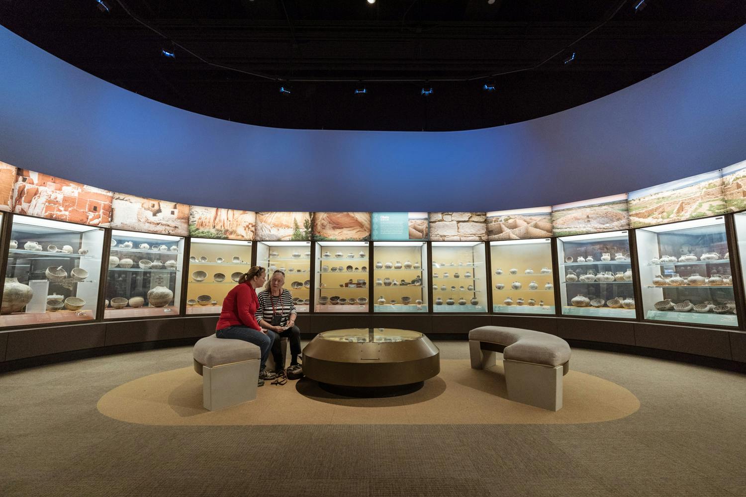Two women sit on a bench in a circular space, surrounded by glass cases filled with pottery in a gallery in Robert R. McCormick Halls of the Ancient Americas.