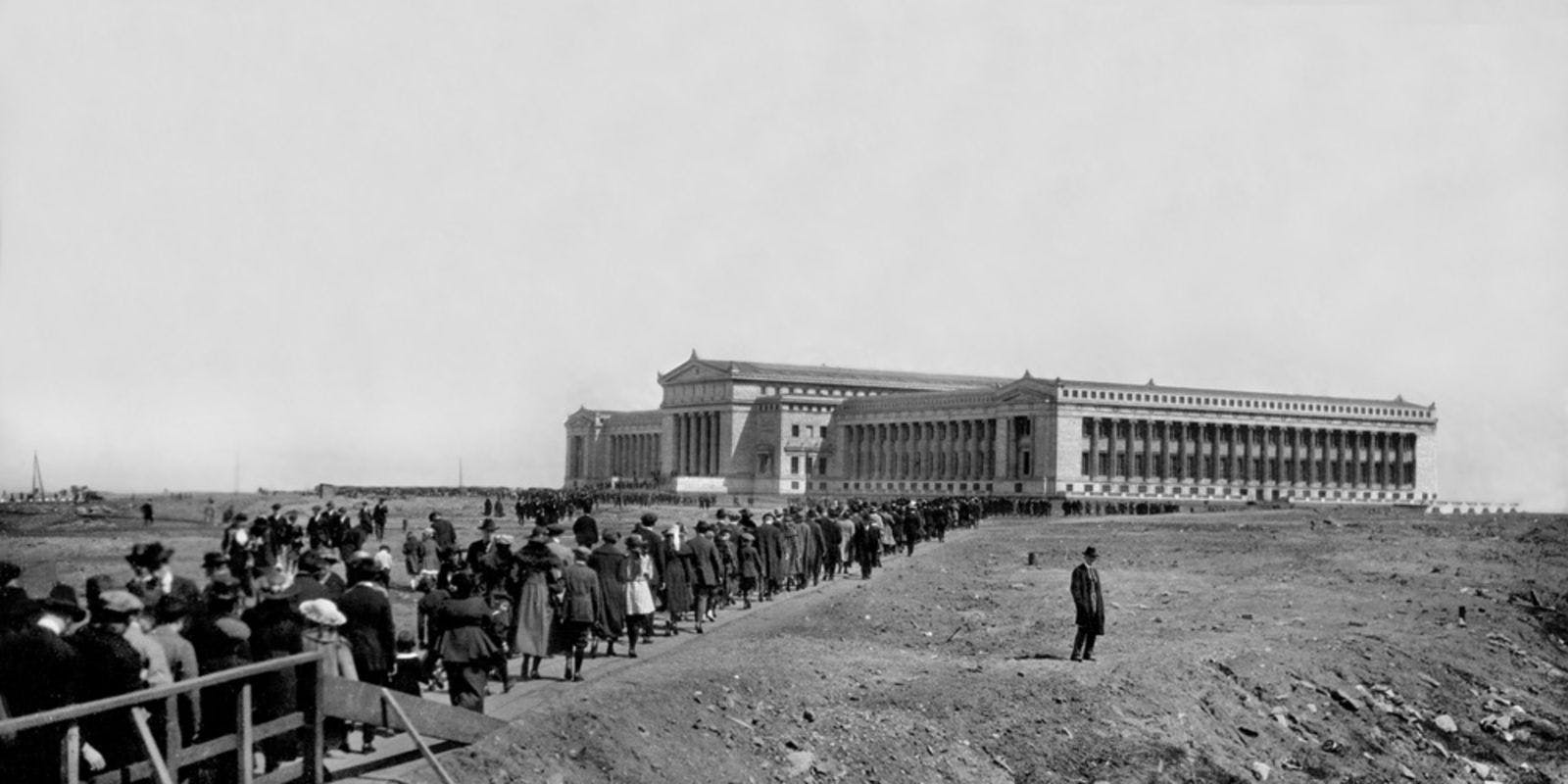 Opening Day of The Field Museum, May 5 1921. Crowds approaching the Museum with a lone man standing to the side of the crowd. People crossing a wooden bridge, with the entire exterior view of the north facade of the Museum in the background.