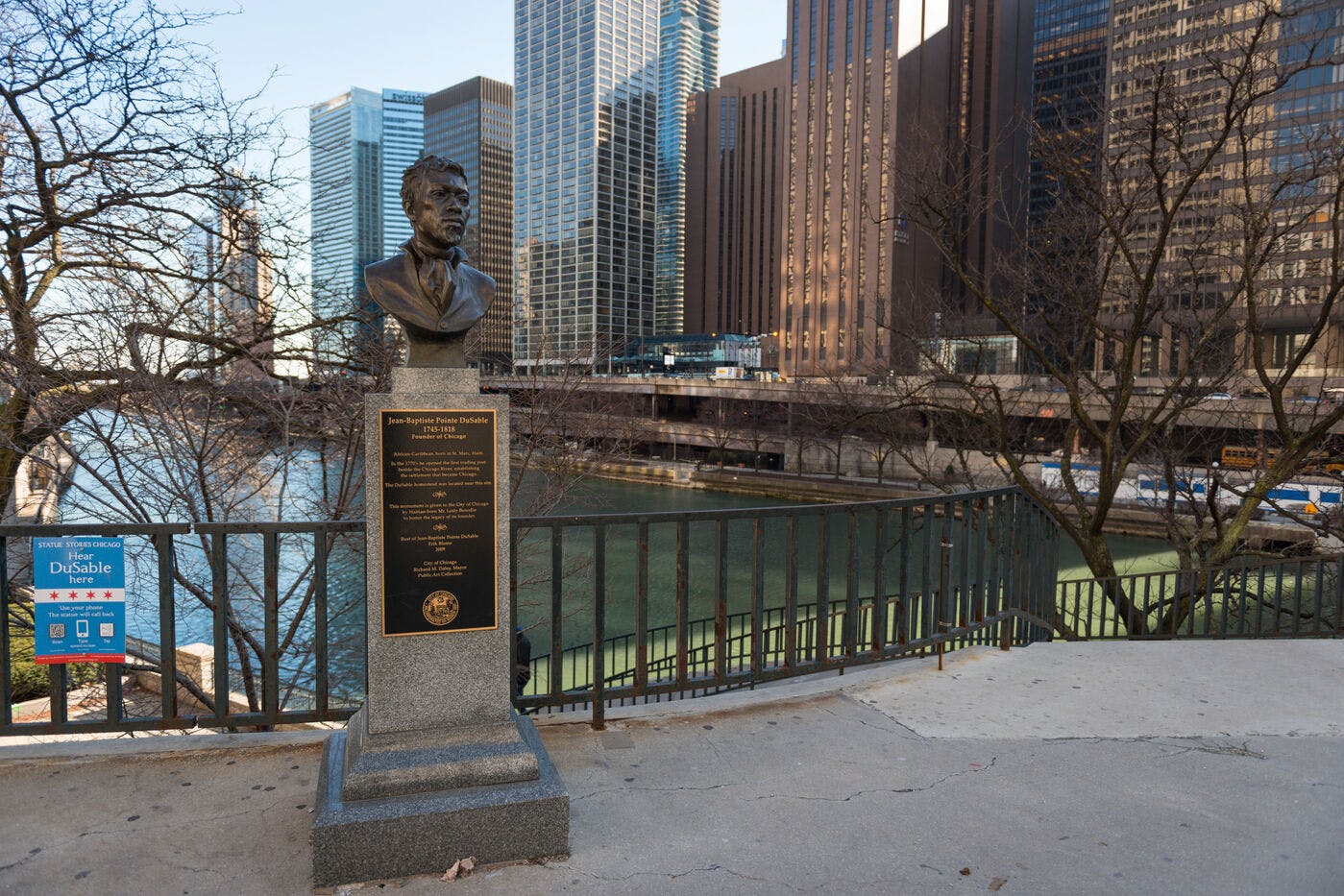 A bronze bust on a pedestal, set on a walkway along the Chicago River with tall buildings in the background. 