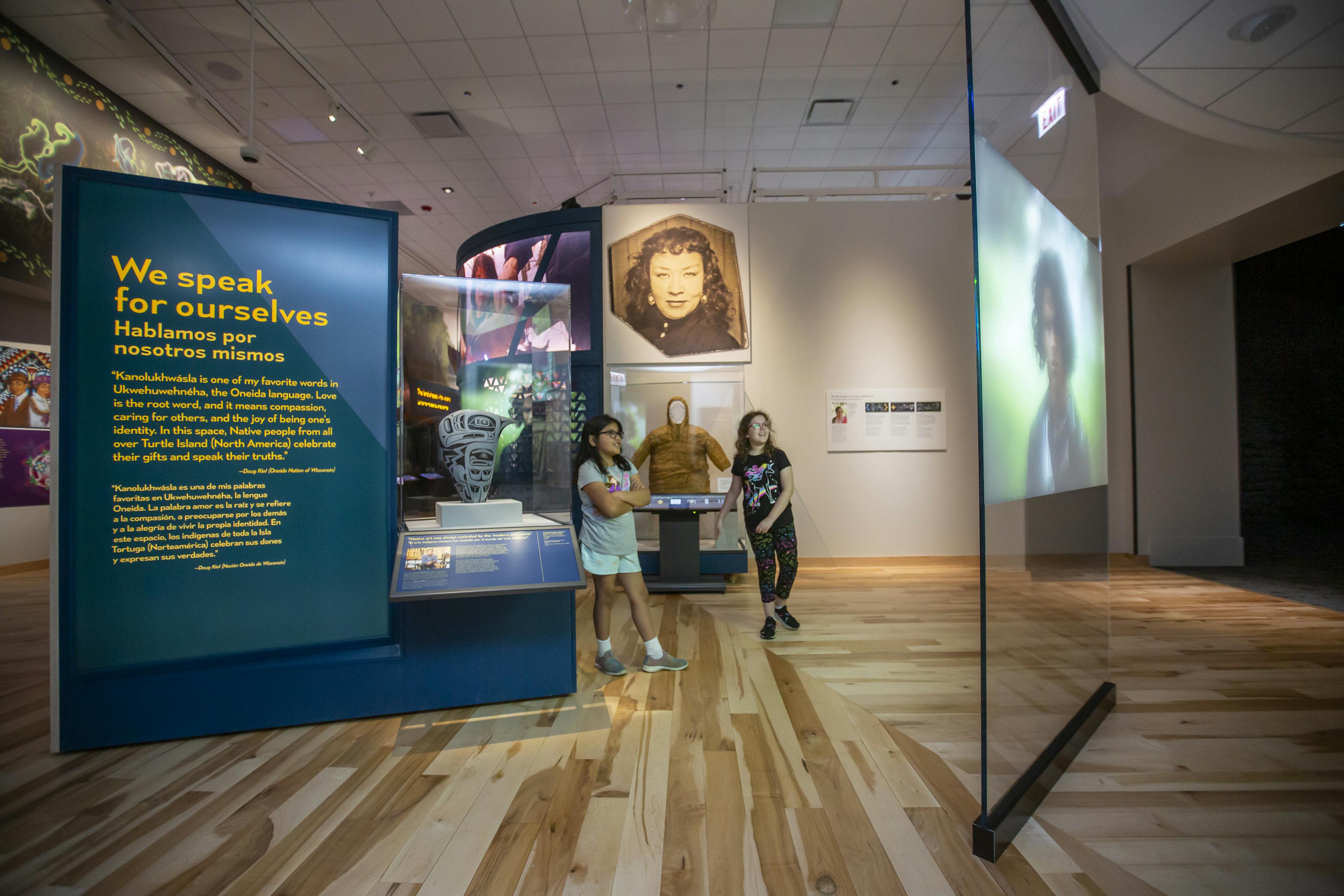 Two young visitors inside the Native Truths exhibition, looking at an interactive floor-to-ceiling screen. Other graphic panels and display cases are behind and next to where they stand.