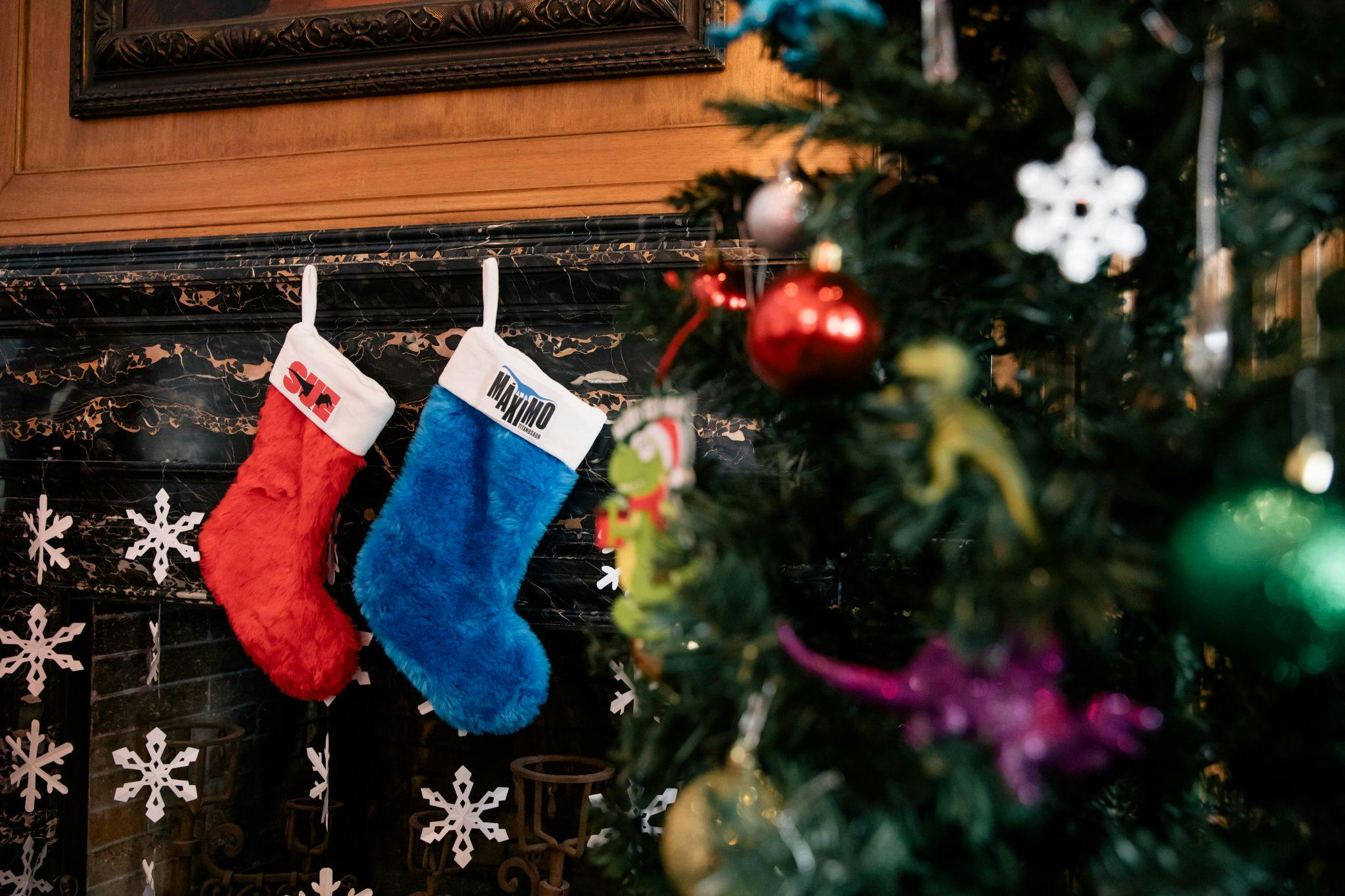 Two Christmas stockings hang from a fireplace mantel– red one with SUE embroidered and a blue one with Maximo embroidered. A Christmas tree is blurred in the foreground to the right.