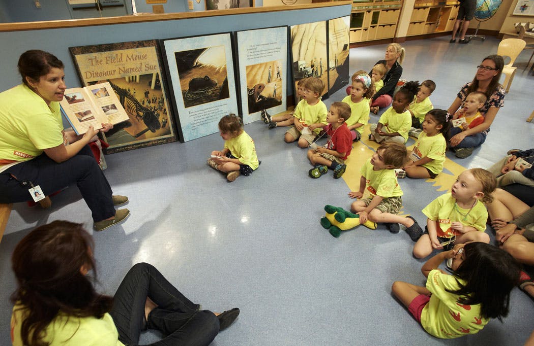 A group of young children sit on the floor listening to a woman read to them. Most everyone in the photo are wearing matching bright yellow t-shirts.