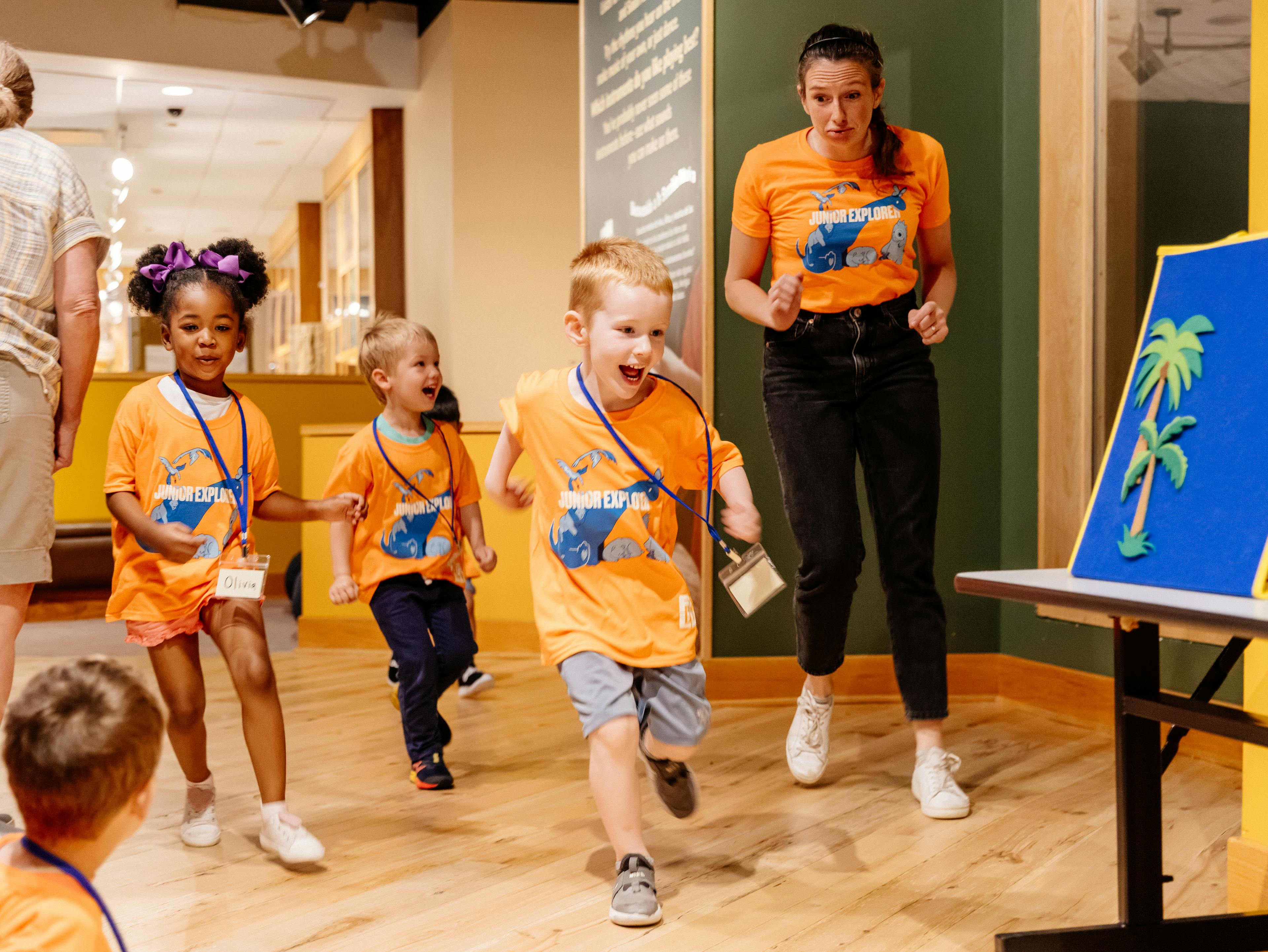 Three children interact with a camp staff member in a museum space. All are wearing matching orange shirts.