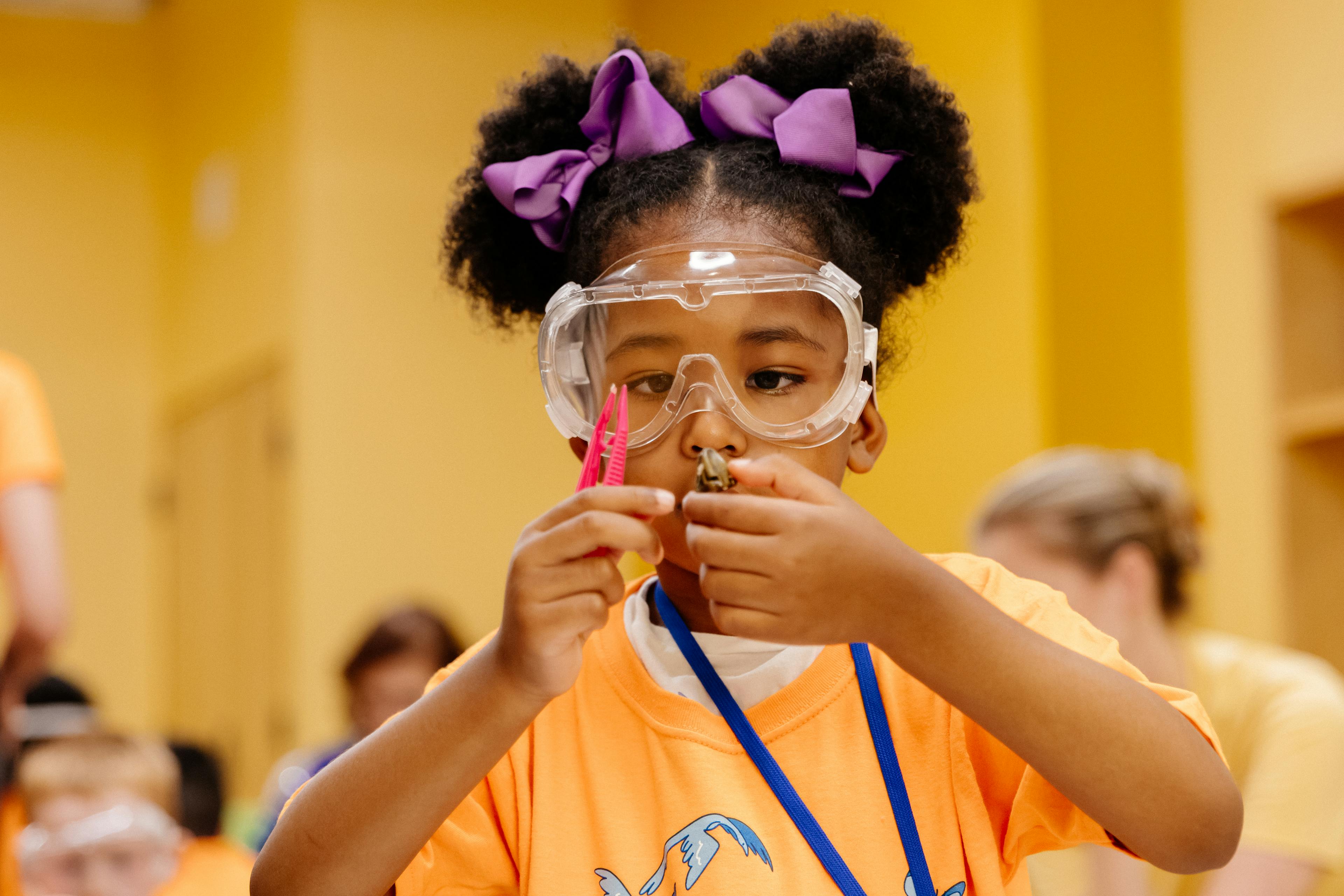 A young child with two buns and purple bows, wearing safety goggles.  The child is holding plastic tweezers and an object that they are holding up looking at through the goggles.
