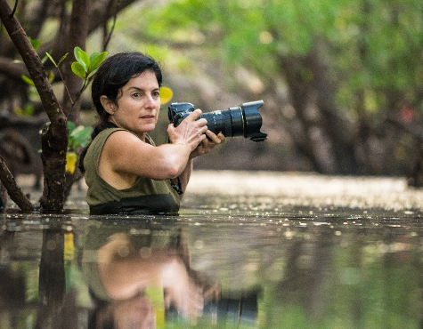 A woman stands in chest deep water, holding a camera with a large lens in both hands.