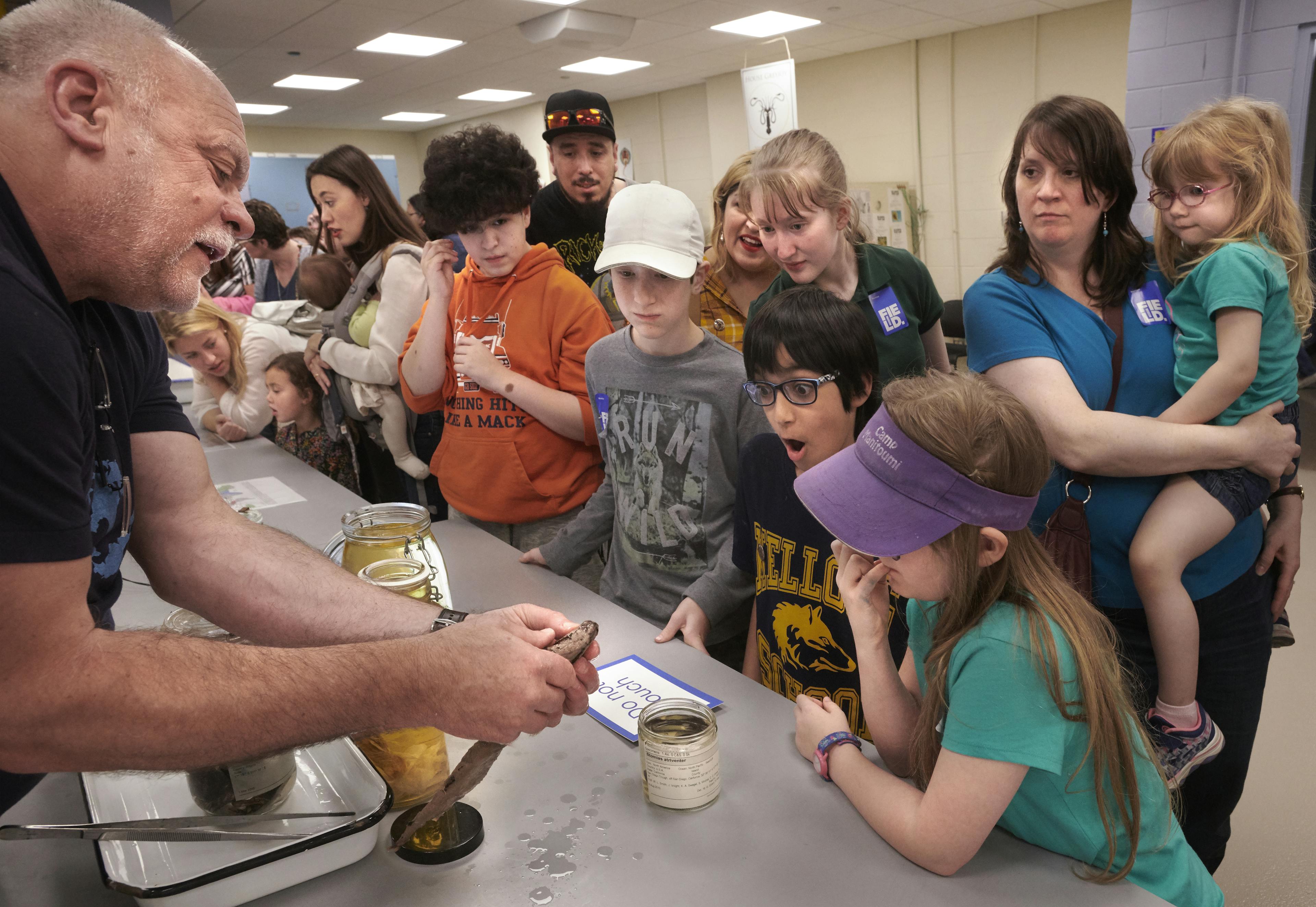 A scientist show a fish specimen to a group of visitors across a table.